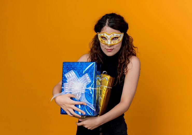 Foto gratuito mujer hermosa joven con el pelo rizado en la máscara del partido que sostiene los regalos que sonríen de pie sobre la pared naranja
