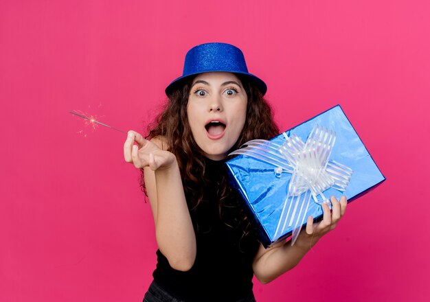 Mujer hermosa joven con el pelo rizado en un sombrero de fiesta con caja de regalo de cumpleaños y bengala concepto de fiesta de cumpleaños sorprendido y asombrado sobre rosa