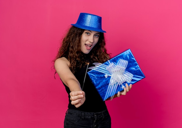 Mujer hermosa joven con el pelo rizado en un sombrero de fiesta con caja de regalo de cumpleaños y bengala feliz y alegre sonriendo alegremente concepto de fiesta de cumpleaños de pie sobre la pared rosa