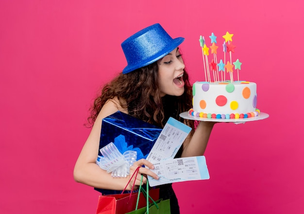 Mujer hermosa joven con el pelo rizado en un sombrero de fiesta con caja de regalo de pastel de cumpleaños y boletos de avión concepto de fiesta de cumpleaños feliz y emocionado de pie sobre la pared rosa