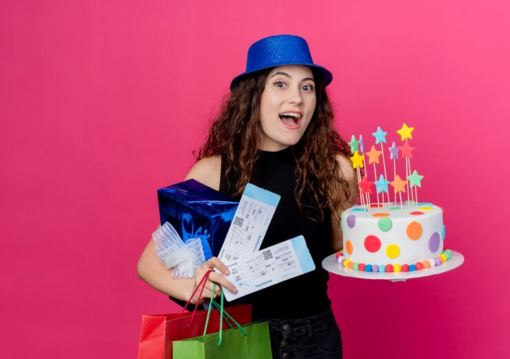 Foto gratuito mujer hermosa joven con el pelo rizado en un sombrero de fiesta con caja de regalo de pastel de cumpleaños y boletos de avión concepto de fiesta de cumpleaños feliz y emocionado de pie sobre la pared rosa