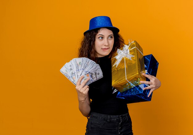 Mujer hermosa joven con el pelo rizado en el sombrero de fiesta con dinero en efectivo y regalos mirando canera feliz y alegre de pie sobre la pared naranja