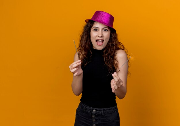 Mujer hermosa joven con el pelo rizado en el sombrero de fiesta con luces de Bengala feliz y emocionado de pie sobre la pared naranja