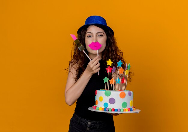 Mujer hermosa joven con el pelo rizado en el sombrero de fiesta con pastel de cumpleaños y palos de fiesta feliz y alegre de pie sobre la pared naranja