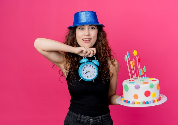 Mujer hermosa joven con el pelo rizado en un sombrero de fiesta con pastel de cumpleaños y reloj despertador mirando sorprendido y feliz cumpleaños concepto de fiesta de pie sobre la pared rosa