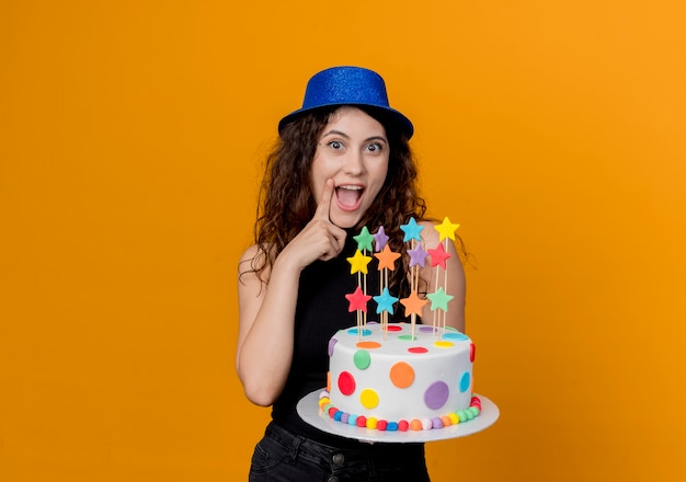 Mujer hermosa joven con el pelo rizado en un sombrero de fiesta sosteniendo la torta de cumpleaños alegre y alegre de pie sobre la pared naranja