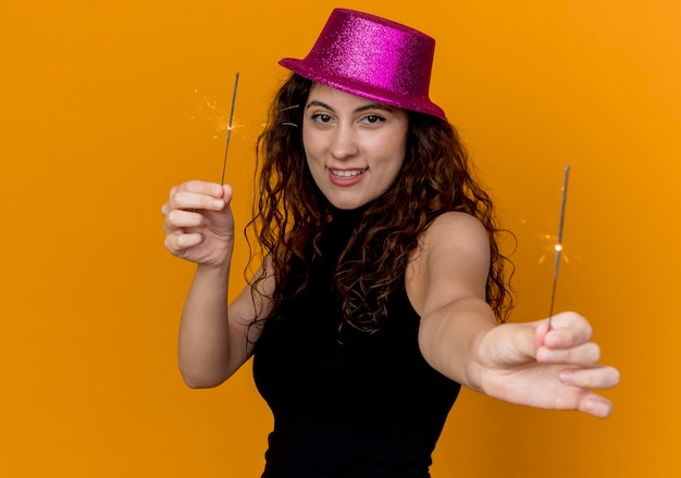 Mujer hermosa joven con el pelo rizado en el sombrero del partido que muestra bengalas feliz y alegre sonriente de pie sobre la pared naranja