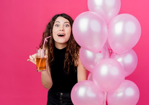 Foto gratuita mujer hermosa joven con el pelo rizado sosteniendo un montón de globos de aire y un cóctel mirando sorprendido sonriendo alegremente de pie sobre la pared rosa