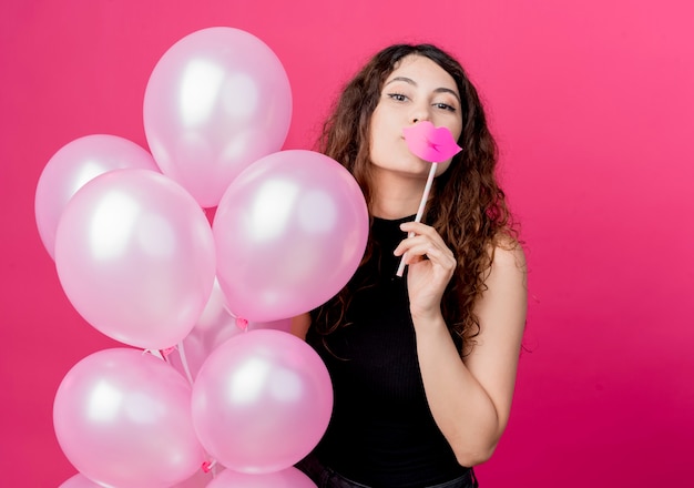 Mujer hermosa joven con el pelo rizado sosteniendo un montón de globos de aire y palo de fiesta sonriendo alegremente de pie sobre la pared rosa