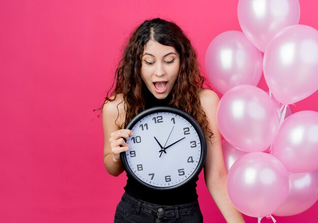 Mujer hermosa joven con el pelo rizado sosteniendo un montón de globos de aire reloj de pared mirándolo sorprendido y asombrado de pie sobre la pared rosa