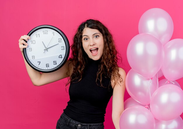 Mujer hermosa joven con el pelo rizado sosteniendo un montón de globos de aire y un reloj de pared sonriendo alegremente de pie sobre la pared rosa