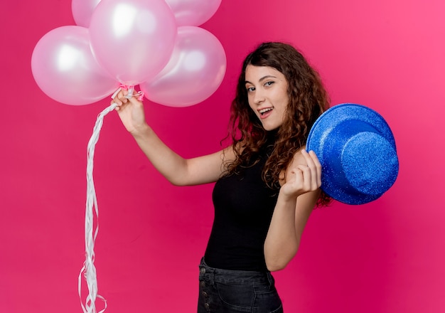 Foto gratuita mujer hermosa joven con el pelo rizado sosteniendo un montón de globos de aire y sombrero de fiesta sonriendo alegremente de pie sobre la pared rosa