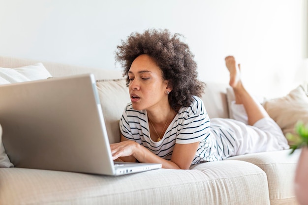 Mujer hermosa joven con el pelo rizado trabajando en la computadora portátil mientras está sentado en el sofá en casa