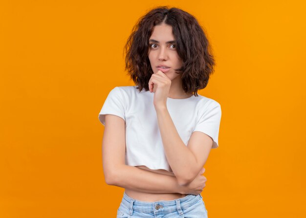 Mujer hermosa joven pensativa que pone la mano en la barbilla en la pared naranja aislada con espacio de copia