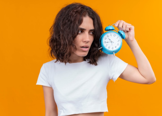 Mujer hermosa joven pensativa que sostiene el reloj de alarma en la pared naranja aislada con espacio de copia