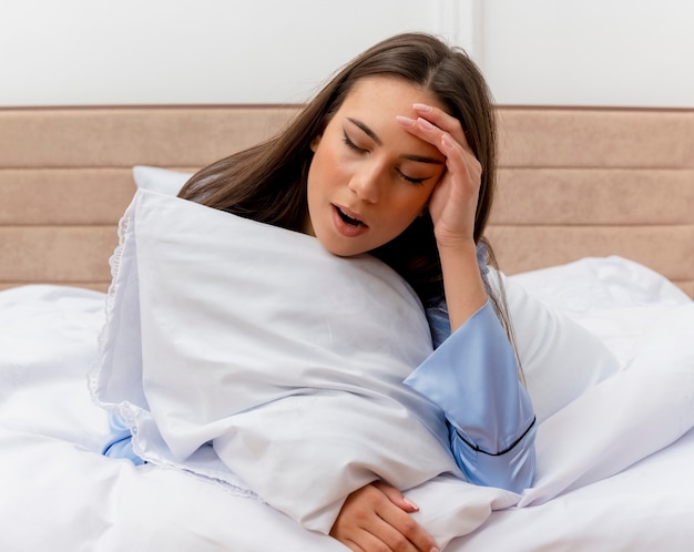 Mujer hermosa joven en pijama azul acostado en la cama con almohada bostezando sensación de fatiga matutina en el interior del dormitorio sobre fondo claro