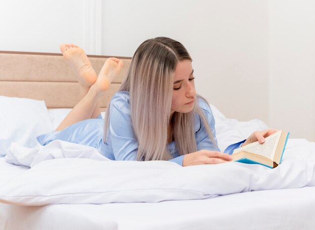 Mujer hermosa joven en pijama azul acostado en la cama descansando libro de lectura en el interior del dormitorio