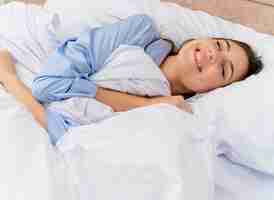 Foto gratuita mujer hermosa joven en pijama azul acostado en la cama descansando sobre suaves almohadas disfrutando de la mañana sonriendo con los ojos cerrados en el interior del dormitorio sobre fondo claro