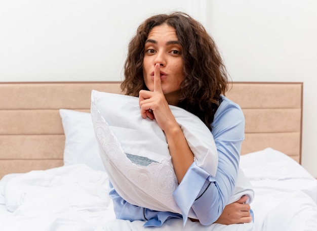 Mujer hermosa joven en pijama azul sentada en la cama con almohada haciendo gesto de silencio con el dedo en los labios en el interior del dormitorio sobre fondo claro