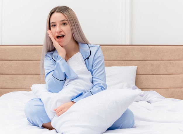 Mujer hermosa joven en pijama azul sentada en la cama con almohada mirando a cámara feliz y sorprendido en el interior del dormitorio