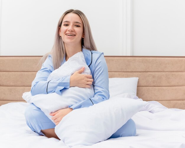 Mujer hermosa joven en pijama azul sentada en la cama con almohada mirando a la cámara sonriendo con cara feliz en el interior del dormitorio sobre fondo claro