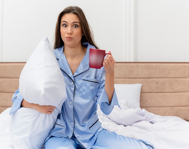 Mujer hermosa joven en pijama azul sentada en la cama con almohada y taza de café mirando a cámara confundida en el interior del dormitorio sobre fondo claro