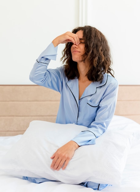 Foto gratuita mujer hermosa joven en pijama azul sentada en la cama con la almohada tocando la nariz entre los ojos cerrados sintiendo fatiga matutina en el interior del dormitorio sobre fondo claro