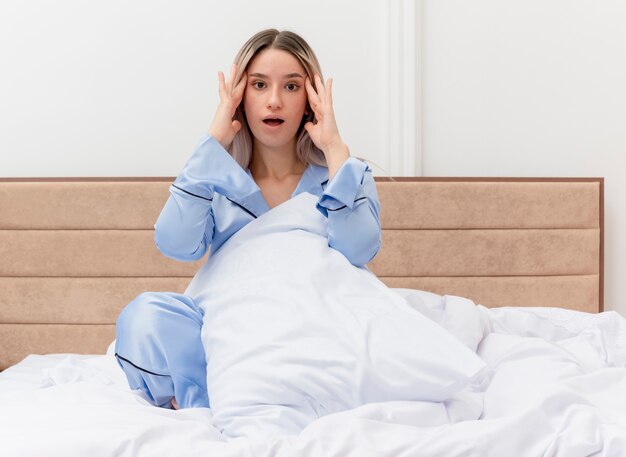 Mujer hermosa joven en pijama azul sentada en la cama con la almohada tocando sus sienes mirando confundido en el interior del dormitorio