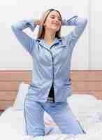 Foto gratuita mujer hermosa joven en pijama azul sentada en la cama descansando sintiendo emociones positivas sonriendo en el interior del dormitorio sobre fondo claro