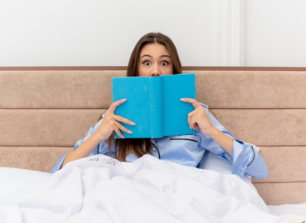 Mujer hermosa joven en pijama azul sentada en la cama con libro mirando a cámara sorprendido en el interior del dormitorio sobre fondo claro