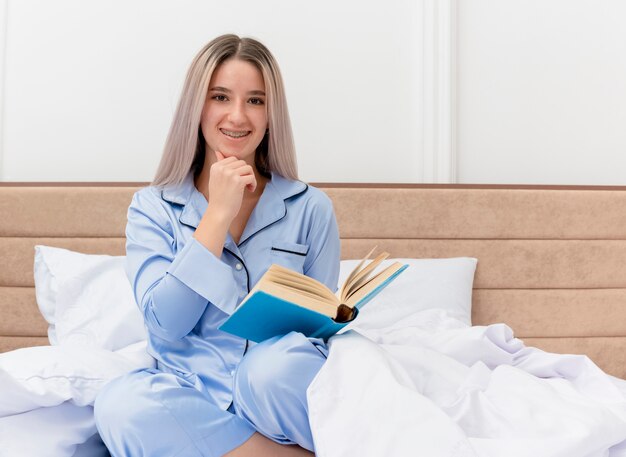 Mujer hermosa joven en pijama azul sentada en la cama con el libro con una sonrisa en la cara en el interior del dormitorio