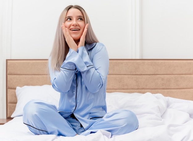 Mujer hermosa joven en pijama azul sentada en la cama mirando asombrado y sorprendido en el interior del dormitorio