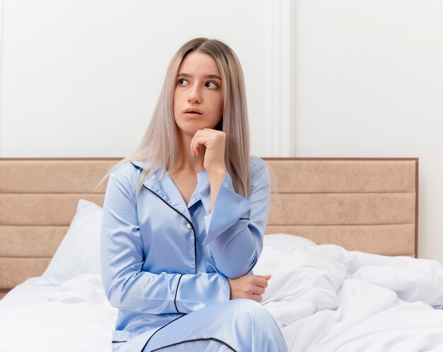 Mujer hermosa joven en pijama azul sentada en la cama mirando a un lado con expresión pensativa en el interior del dormitorio sobre fondo claro