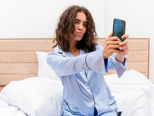 Mujer hermosa joven en pijama azul sentada en la cama con smartphone haciendo selfie soplando un beso en el interior del dormitorio sobre fondo claro