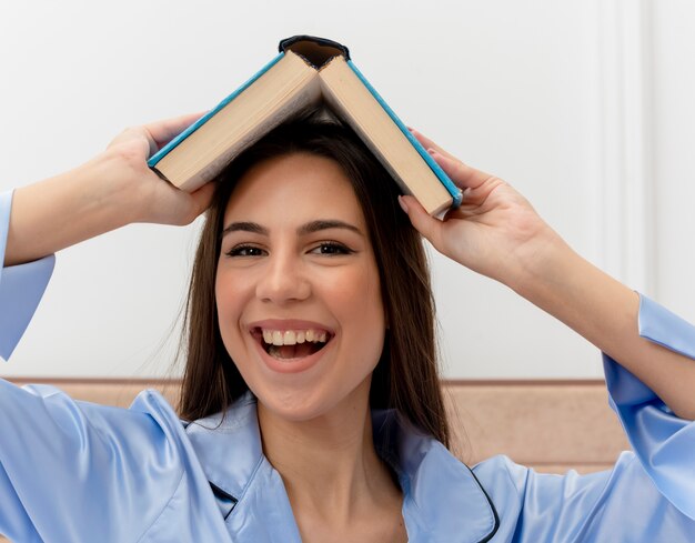 Mujer hermosa joven en pijama azul sentada en la cama sosteniendo el libro sobre su cabeza mirando alegre feliz y alegre sonriendo en el interior del dormitorio sobre fondo claro