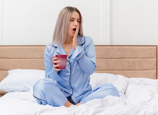 Mujer hermosa joven en pijama azul sentada en la cama con una taza de café bostezando despertarse sintiendo fatiga matutina en el interior del dormitorio