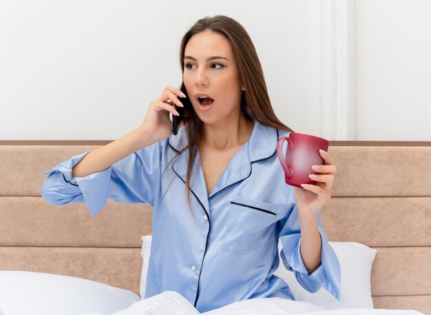 Mujer hermosa joven en pijama azul sentada en la cama con una taza de café hablando por teléfono móvil que se sorprende en el interior del dormitorio sobre fondo claro