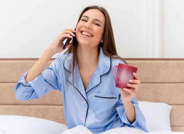 Mujer hermosa joven en pijama azul sentada en la cama con una taza de café hablando por teléfono móvil sonriendo alegremente en el interior del dormitorio sobre fondo claro