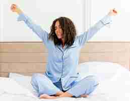 Foto gratuita mujer hermosa joven en pijama azul sentado en la cama estirando las manos despertando feliz y positivo disfrutando de la mañana en el interior del dormitorio sobre fondo claro