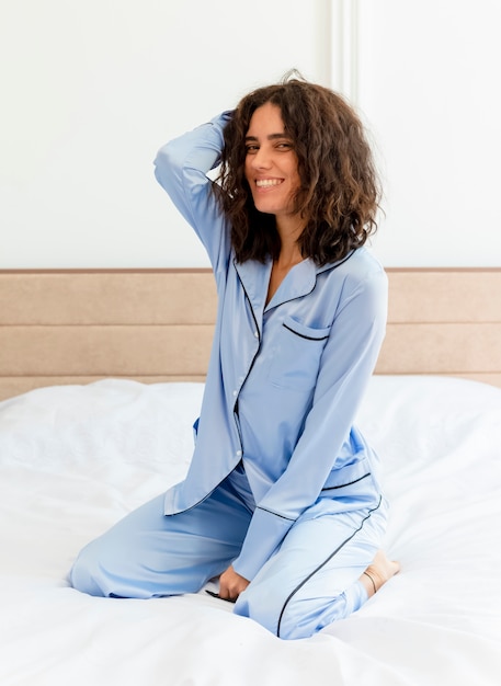 Mujer hermosa joven en pijama azul sentado en la cama mirando a cámara con una sonrisa en la cara feliz y positiva en el interior del dormitorio sobre fondo claro