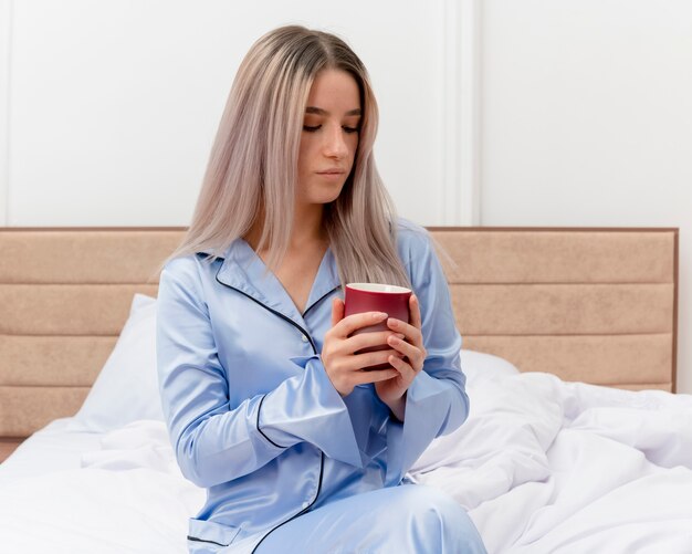 Mujer hermosa joven en pijama azul sentado en la cama con una taza de café mirando hacia abajo pensando en el interior del dormitorio sobre fondo claro