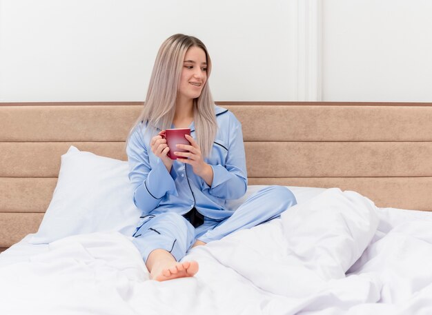 Mujer hermosa joven en pijama azul sentado en la cama con una taza de café mirando a un lado sonriendo descansando disfrutando del tiempo de la mañana en el interior del dormitorio