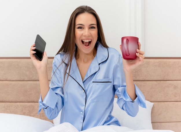 Mujer hermosa joven en pijama azul sentado en la cama con una taza de café con smartphone mirando a la cámara feliz y emocionado en el interior del dormitorio sobre fondo claro