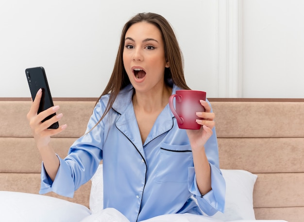 Mujer hermosa joven en pijama azul sentado en la cama con una taza de café con smartphone sorprendido y sorprendido en el interior del dormitorio sobre fondo claro