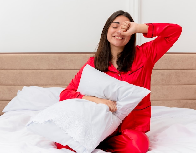 Mujer hermosa joven en pijama rojo sentada en la cama con almohada quiere dormir bostezando en el interior del dormitorio sobre fondo claro