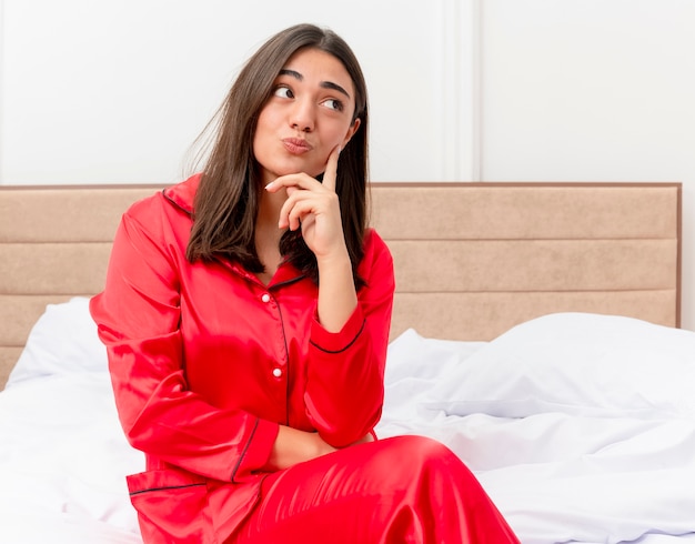 Mujer hermosa joven en pijama rojo sentada en la cama mirando hacia arriba con expresión pensinve en la cara pensando en el interior del dormitorio sobre fondo claro