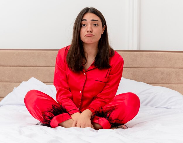 Mujer hermosa joven en pijama rojo sentada en la cama mirando a cámara con expresión triste en la cara en el interior del dormitorio sobre fondo claro