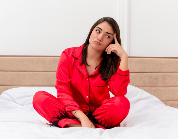 Mujer hermosa joven en pijama rojo sentada en la cama mirando a un lado con expresión pensativa disgustada en el interior del dormitorio sobre fondo claro