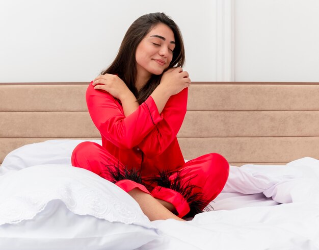 Mujer hermosa joven en pijama rojo sentada en la cama con los ojos cerrados abrazándose a sí misma sintiendo emociones positivas en el interior del dormitorio