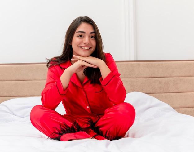 Foto gratuita mujer hermosa joven en pijama rojo sentada en la cama sonriendo con cara feliz posando a la cámara con la cabeza apoyada en las palmas en el interior del dormitorio sobre fondo claro